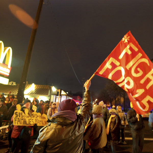 Demonstration framför ett mcdonalds. Det är kväll och mörkt och en demonstrant håller en röd flagga med texten Fight for 15. McDonalds-M:et syns i bakgrunden.