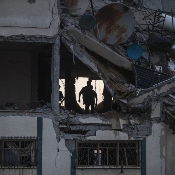 Bombat hus med hål i fasaden och infallet tak. Inne i huset syns människor som söker efter överlevande.